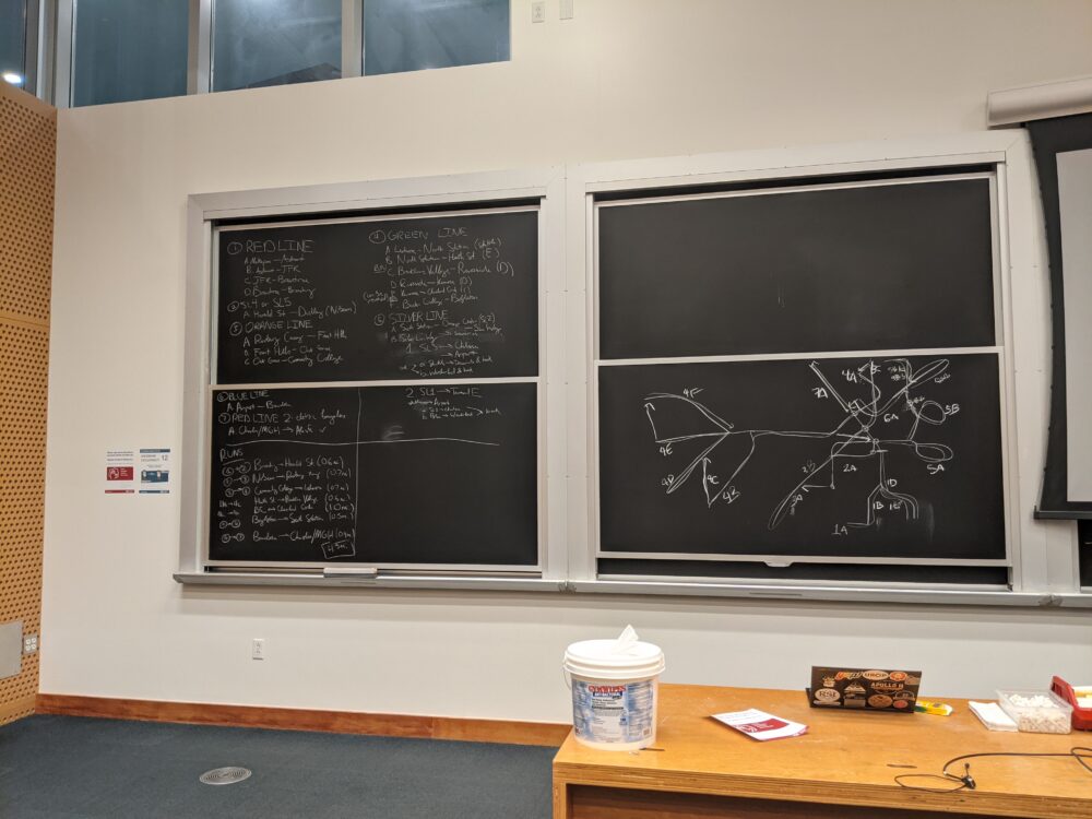 two sets of blackboards in a classroom: the left is filled with text describing a route, the right has a diagram of the MBTA map drawn with arrows and annotations