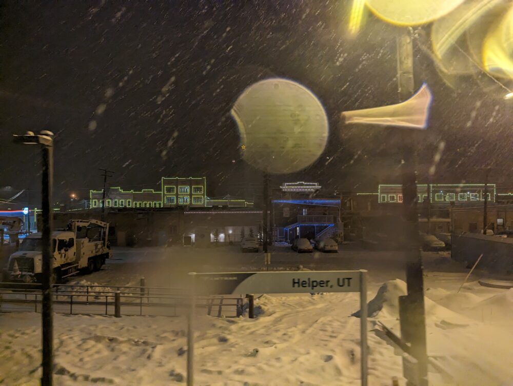 image of amtrak sign reading Helper, UT, in thick snowstorm with buildings with Christmas lights in the background